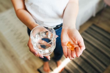 Close up of person holding vitamin capsules in one hand and a glass of water in the other, showing taking vitamins before or after a meal